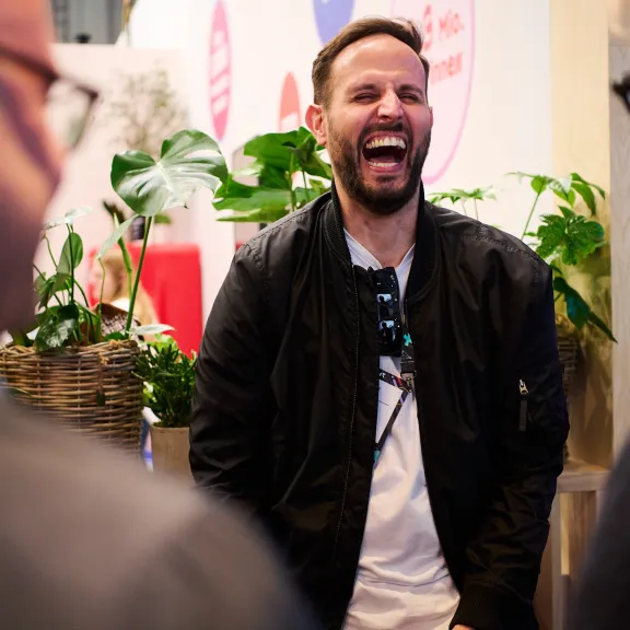 Mann lacht herzlich im Gespräch mit zwei anderen Personen, Pflanzen im Hintergrund.
