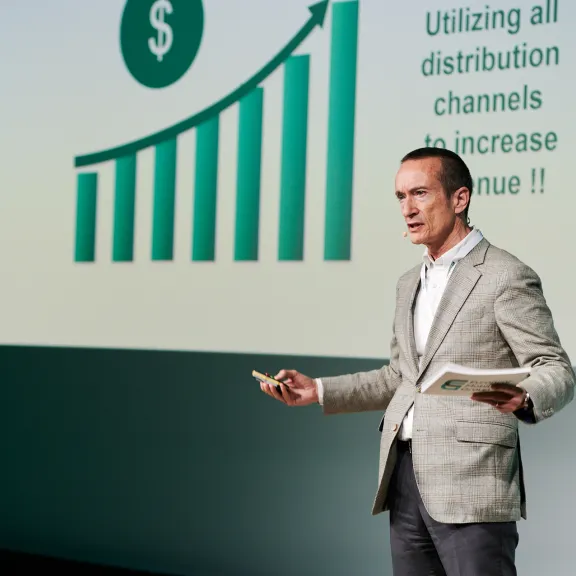 Mann im Anzug präsentiert mit Diagramm und Text auf Leinwand im Hintergrund.