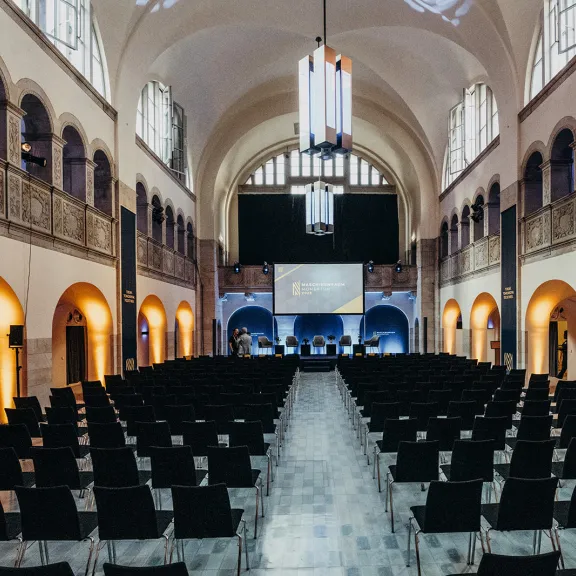 Großer Saal mit leeren Stuhlreihen, Bühne und hohen Decken, festlich beleuchtet.