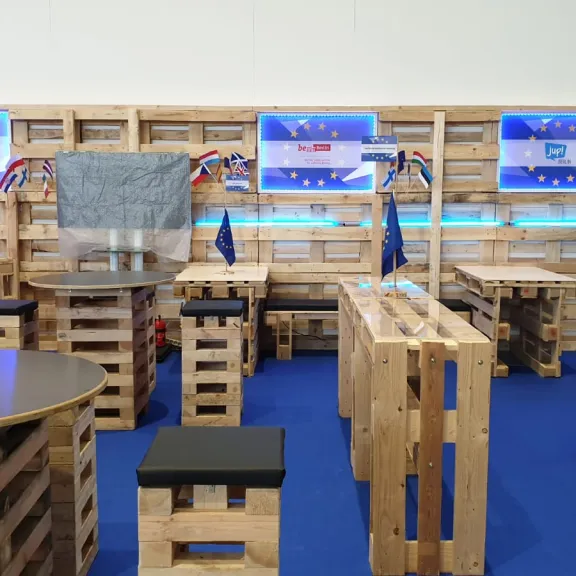 Aus Holzpaletten gebaute Sitzmöbel, europäische Fahnen, blaue Teppiche und Bildschirme.