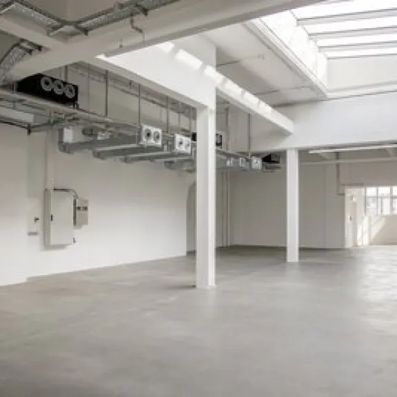 Leere, helle Industriehalle mit Betonboden und großen Fenstern.