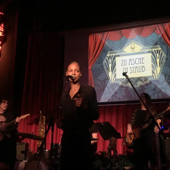 Sänger und Band auf Bühne mit rotem Vorhang und beleuchtetem "So Fackin Wild"-Schild im Hintergrund