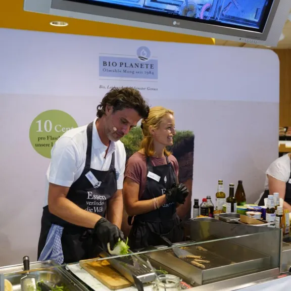 Zwei Personen kochen gemeinsam an einem Messestand mit Kochutensilien und Zutaten.