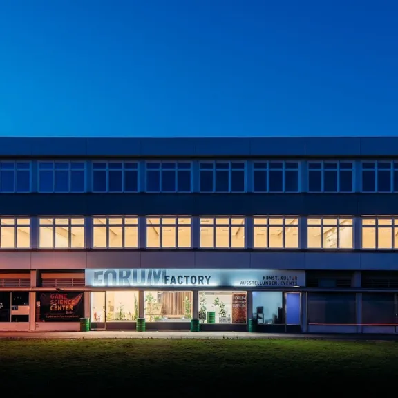Forum Factory Berlin bei Abenddämmerung: Das Gebäude mit Grünfläche im industriellen Gewerbestil mit beleuchteten Fenstern.