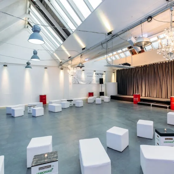 Heller Veranstaltungsraum mit weißen Sitzwürfeln und Bühne, große Fenster durch das Sheddach und Vorhänge.