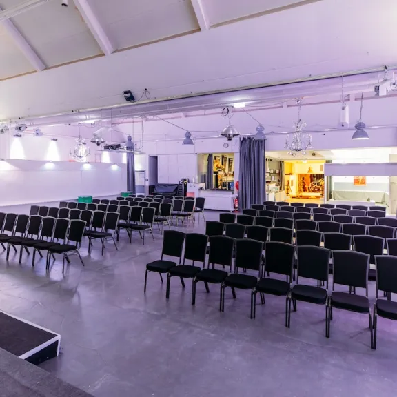 Heller Veranstaltungsraum der Forum Factory Berlin mit atmosphärischer Beleuchtung. Stühle für ca. 100 Personen stehen in Reihe vor der Bühne