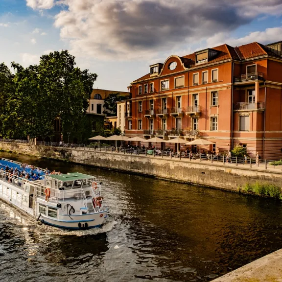 Weiße Ausflugsboot fährt an rotem Backsteingebäude auf der Spree entlang.