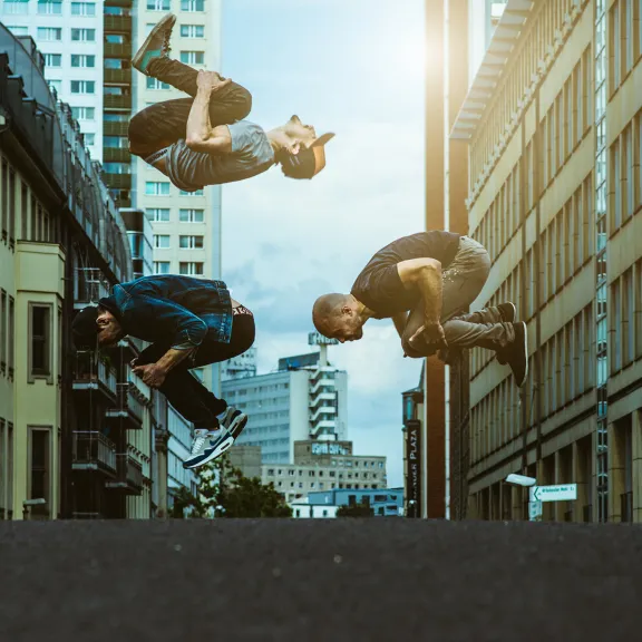 Drei Männer machen Rückwärtssaltos auf einer Straße zwischen Hochhäusern.
