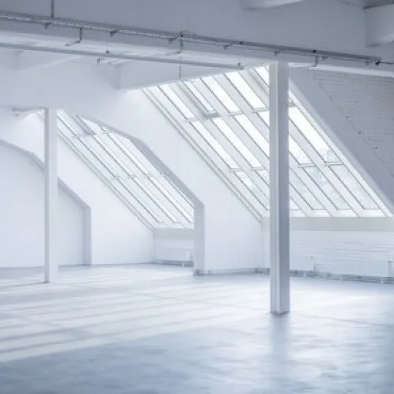 Großer, leerer Raum mit schrägen Fenstern und viel Tageslicht