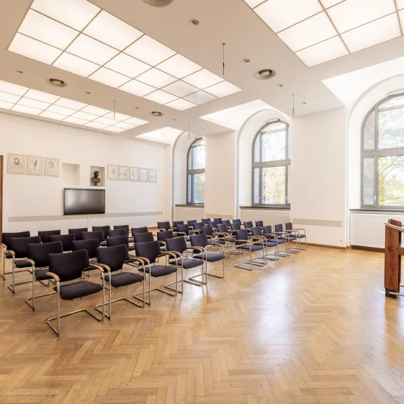 Heller, leerer Konferenzraum mit Stuhlreihen und Rednerpult, große Fenster und Holzboden.