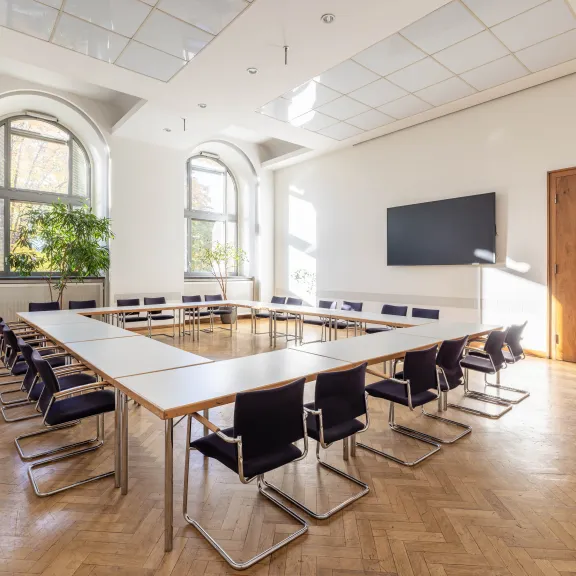 Heller Konferenzraum mit U-förmigem Tisch, Stühlen und großem Bildschirm an der Wand.