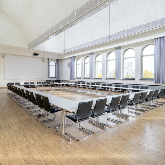 Großer, heller Konferenzraum mit U-förmig angeordneten Tischen und vielen Stühlen.