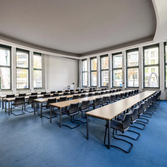 Heller, leerer Seminarraum mit Tischen und Stühlen, große Fenster, blaue Teppiche.