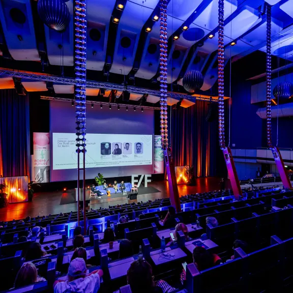 Modernes Auditorium mit Bühne, beleuchtet in Blau und Orange, Publikum schaut zu.