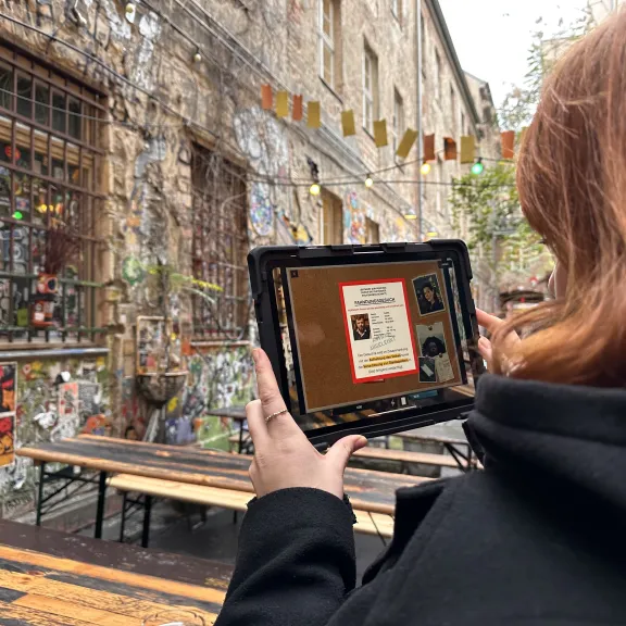 Person hält Tablet vor Grafitti-bedeckter Wand im Außenbereich mit Holzbänken, zeigt Outdoor Escape Game Inside im Einsatz