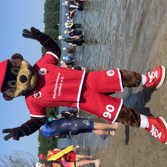 Maskottchen im roten Sportoutfit steht am Seeufer, Menschen im Wasser im Hintergrund.