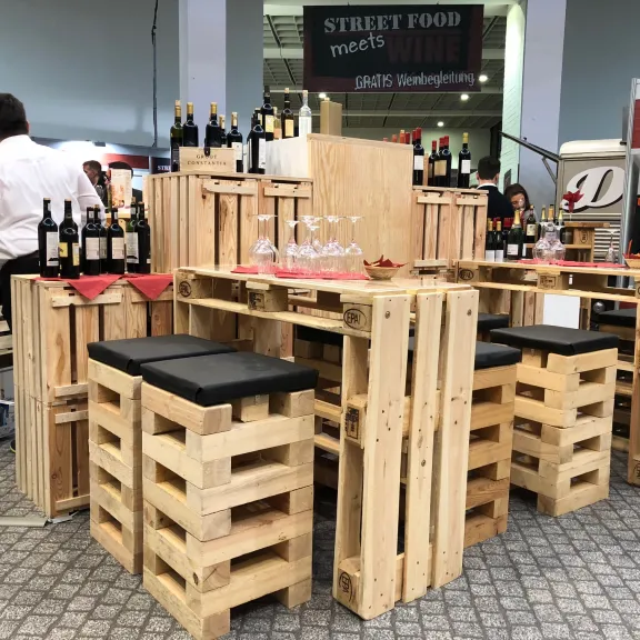 Bar mit Sitzmöbeln aus Paletten, Weinflaschen auf dem Tresen, moderne Messeatmosphäre.