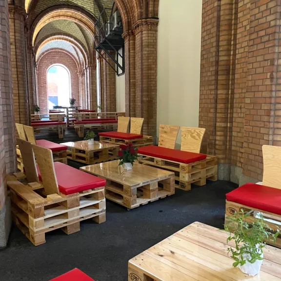 Holzbänke mit roten Polstern und Tischen in einem Backsteinbogen-Gang, Pflanzen auf den Tischen.