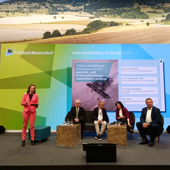 Podiumsdiskussion mit fünf Personen auf einer Bühne, Hintergrund zeigt Felder und Natur.