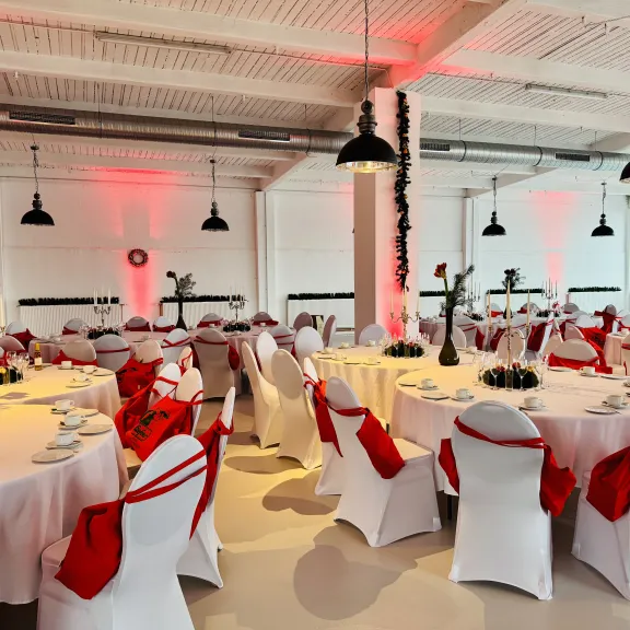 Rund gedeckte Tische mit weißen Hussen und roten Schleifen, festlich dekorierter Saal