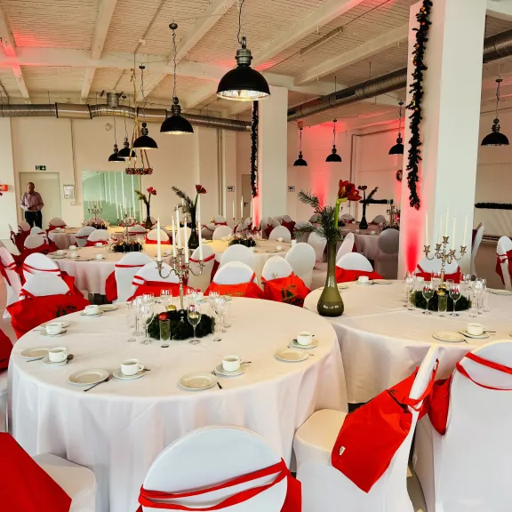 Festlich gedeckter Saal mit runden Tischen, weißen Tischdecken und roten Akzenten.