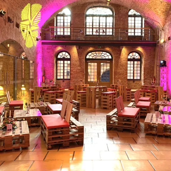 Gemütliche Lounge mit Palettenmöbeln und roten Kissen, Backsteinwände, pinke Beleuchtung.