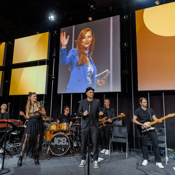 Band mit sechs Mitgliedern spielt auf Bühne, große Bildschirme im Hintergrund.