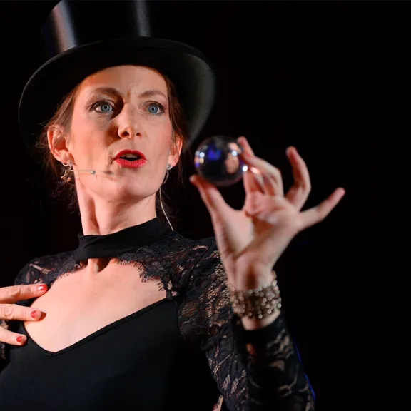 Frau mit Zylinder und schwarzem Kleid hält eine Kugel in der Hand auf dunklem Hintergrund.