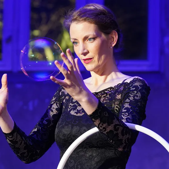 Frau in schwarzem Kleid hält Seifenblase in der Luft, lila Hintergrund