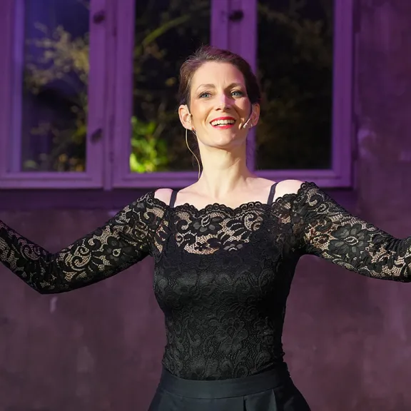 Frau im schwarzen Spitzenkleid lächelt und gestikuliert vor lila beleuchteter Wand.