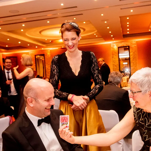 Elegant gekleidete Menschen sitzen lachend an einem festlich gedeckten Tisch im Ballsaal.