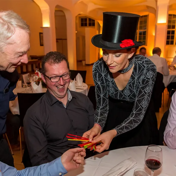 Frau mit Zylinder zeigt Magie-Trick an einem Tisch mit lachenden Männern.