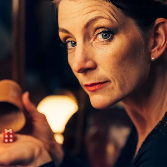 Frau hält einen Würfelbecher und zeigt rote Würfel in ihrer Hand, schaut ernst.