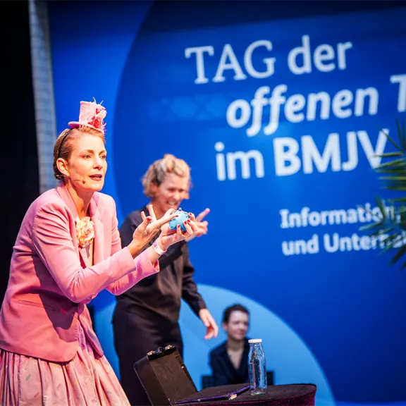 Frau in pinkem Kostüm gestikuliert vor blauem Banner beim Tag der offenen Tür im BMJV.