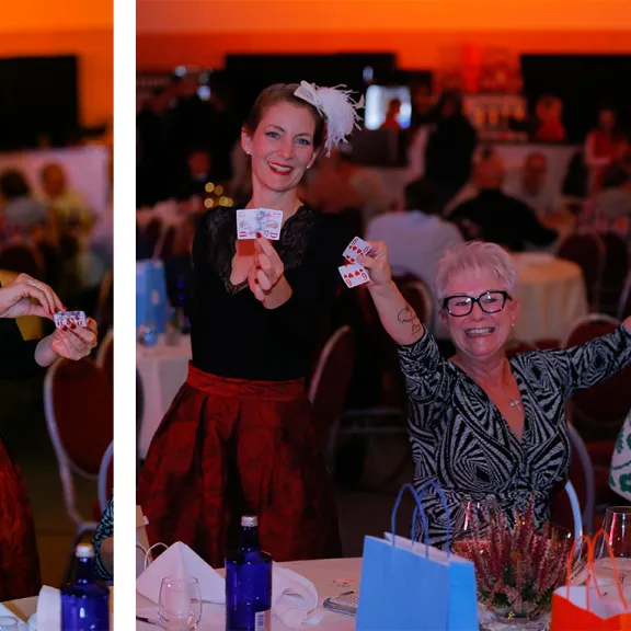 Frau zaubert mit Spielkarte, andere Frauen am Tisch freuen sich und jubeln bei einer Tombola.