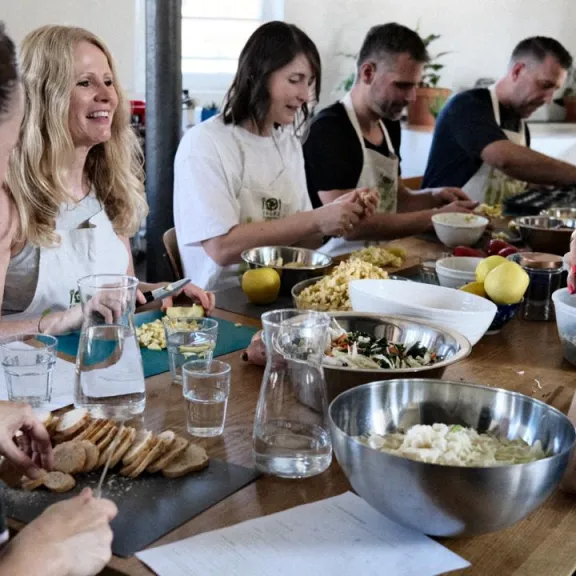 Sechs Menschen bereiten gemeinsam Essen an einem Holztisch in heller Küche zu, lachend und entspannt.