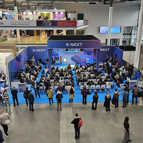 Viele Menschen sitzen und stehen vor einer Bühne auf einer Messe.
