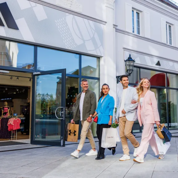 Vier Personen mit Einkaufstaschen gehen lachend vor einem Bekleidungsgeschäft entlang.