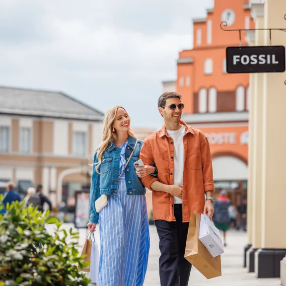Paar spaziert lächelnd mit Einkaufstaschen durch eine Einkaufsstraße bei bewölktem Himmel.