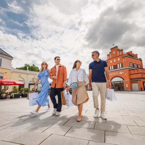 Vier Personen gehen lachend mit Einkaufstaschen durch ein sonniges Einkaufsviertel.