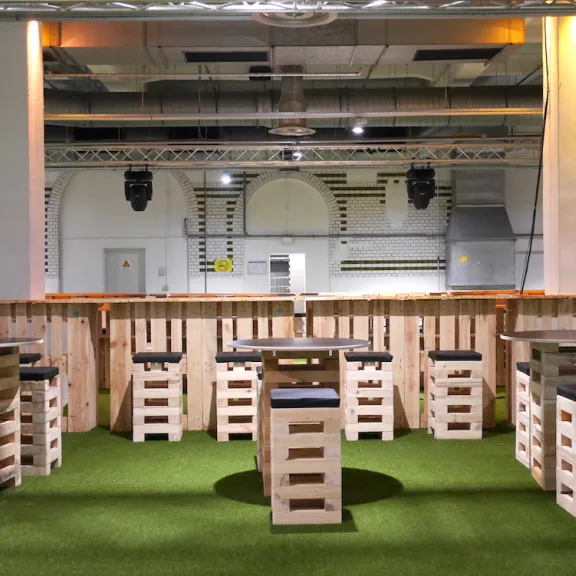 Barbereich mit Holztischen und Hockern auf grünem Teppich, industrielle Decke.