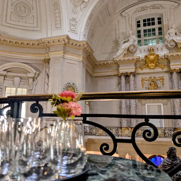 Mehrere leere Gläser und rosa Blumen vor prunkvoller, heller Saaldecke.