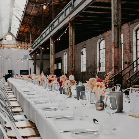 Lange festlich gedeckte Tafel in einer industriellen Halle mit Blumen und Geschirr.