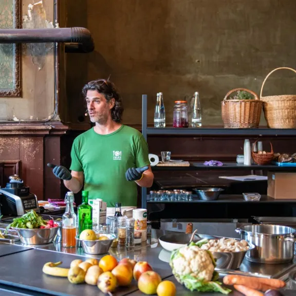 Mann in grüner Kleidung steht in einer Küche mit vielen frischen Zutaten auf dem Tisch.