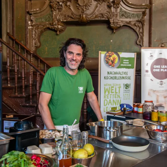 Mann mit langen Haaren in grüner Küche, lächelnd hinter einer Kochstation mit Zutaten.