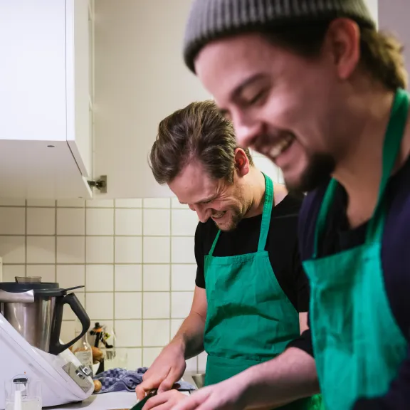 Zwei lachende Männer in grünen Schürzen schneiden Gemüse in einer Küche.
