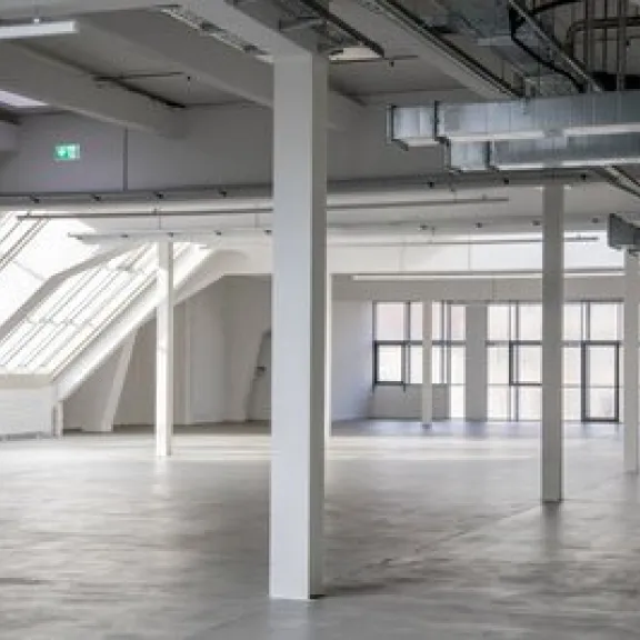 Leere, helle Industriehalle mit Betonboden und sichtbaren Lüftungsrohren.