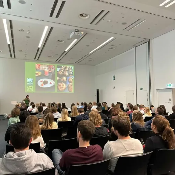 Menschen sitzen in einem modernen Vortragssaal und sehen eine Präsentation auf Leinwand.