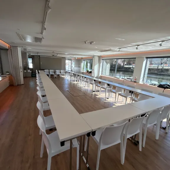 Heller Konferenzraum mit U-förmig angeordneten Tischen und weißen Stühlen.