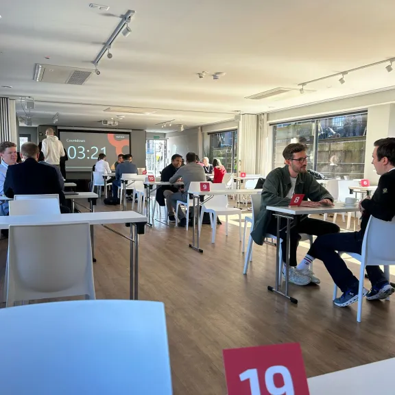 Menschen sitzen an Tischen in einem modernen, hellen Raum; Tischkarte mit Nummer 19 im Vordergrund.